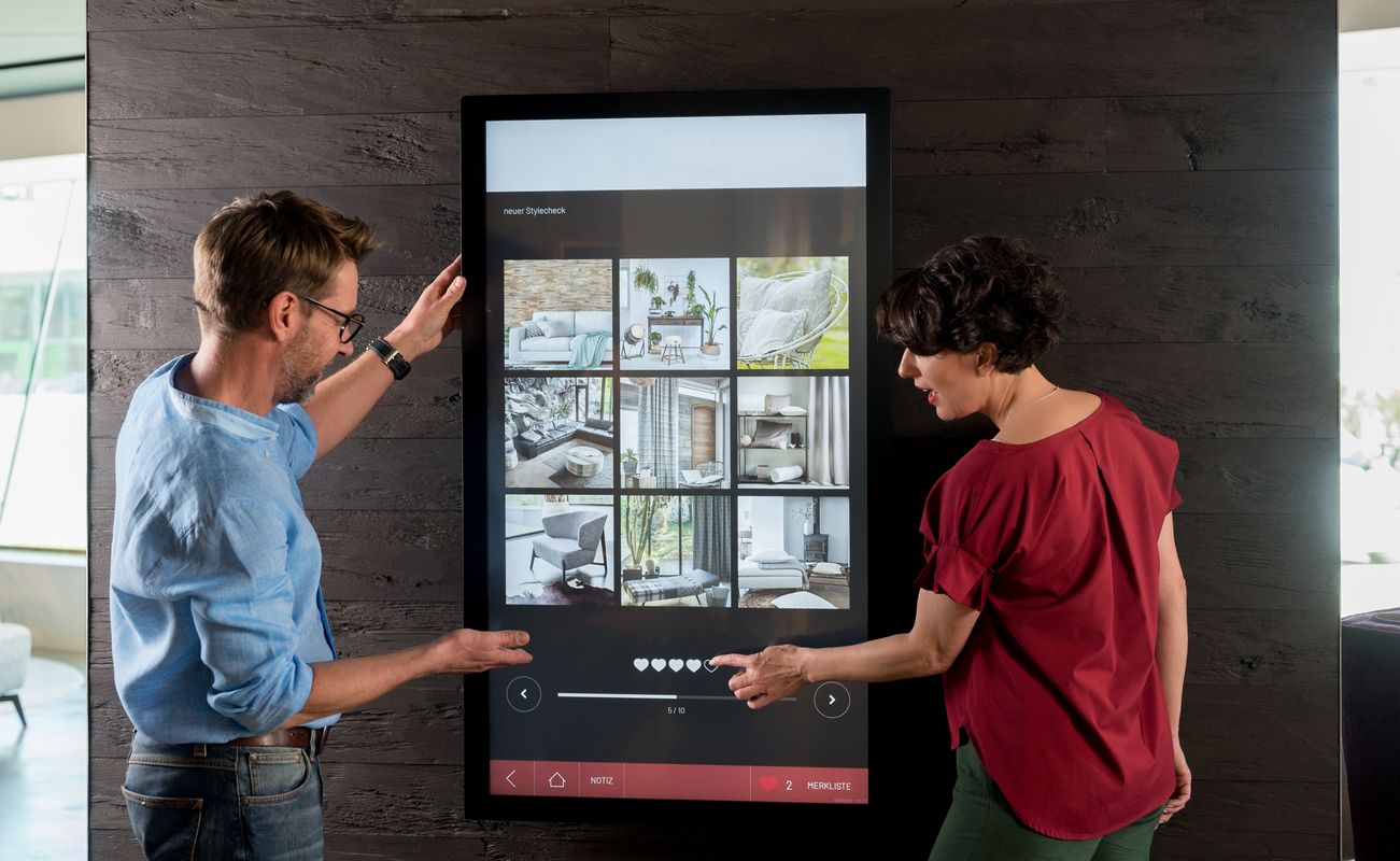 Zwei Personen interagieren mit einem großen Touchscreen, der verschiedene Wohnraumdesigns zeigt. Der Bildschirm ist in einem modernen Raum platziert.